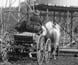 Photograph of a man driving a horse-drawn buggy past a windmill and water tank.