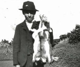 Photograph of a hunter holding up a brace of rabbits he has caught.
